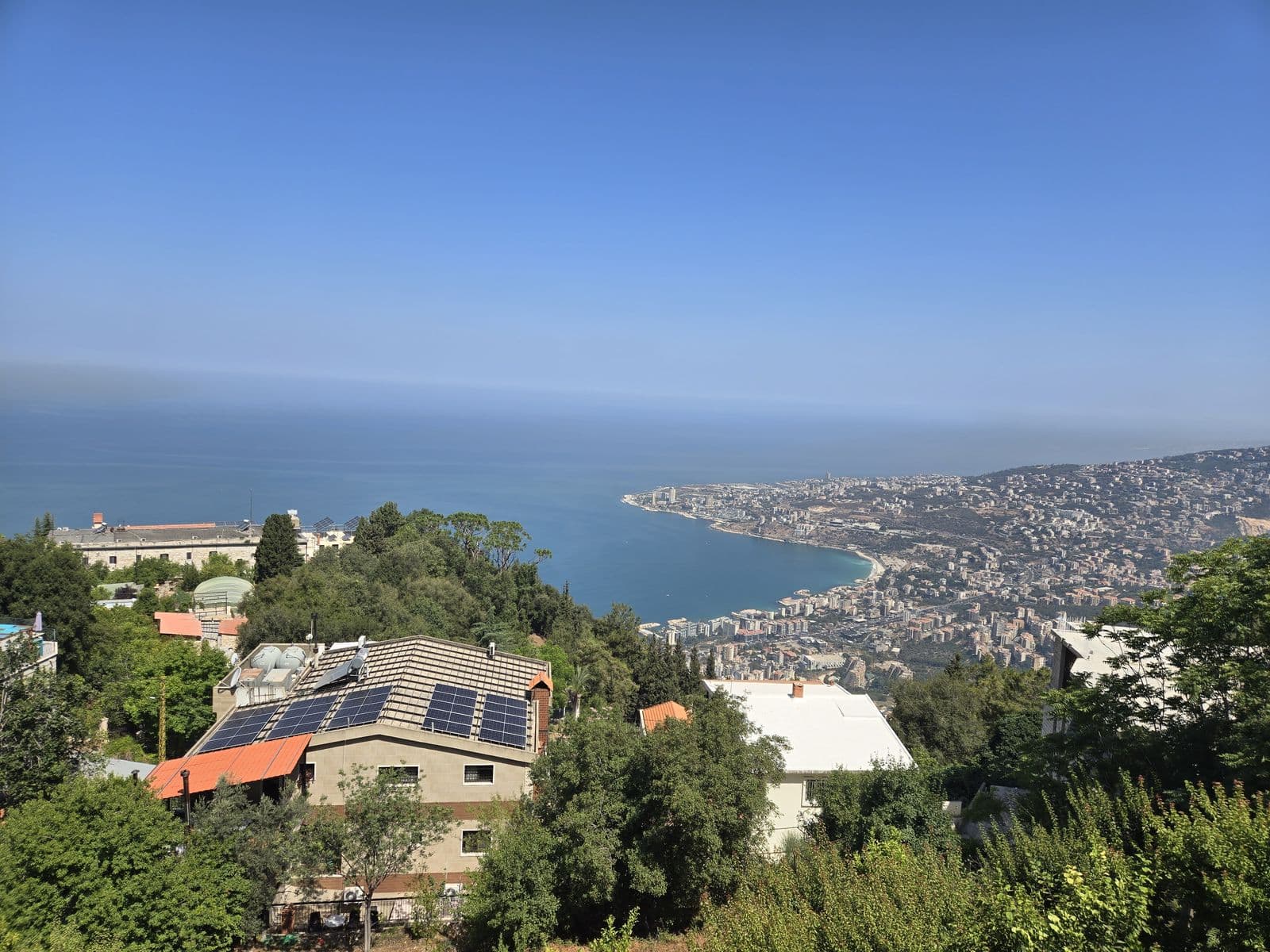 View from Harissa over the bay and the coast, with solar panels on a roof in the foreground.